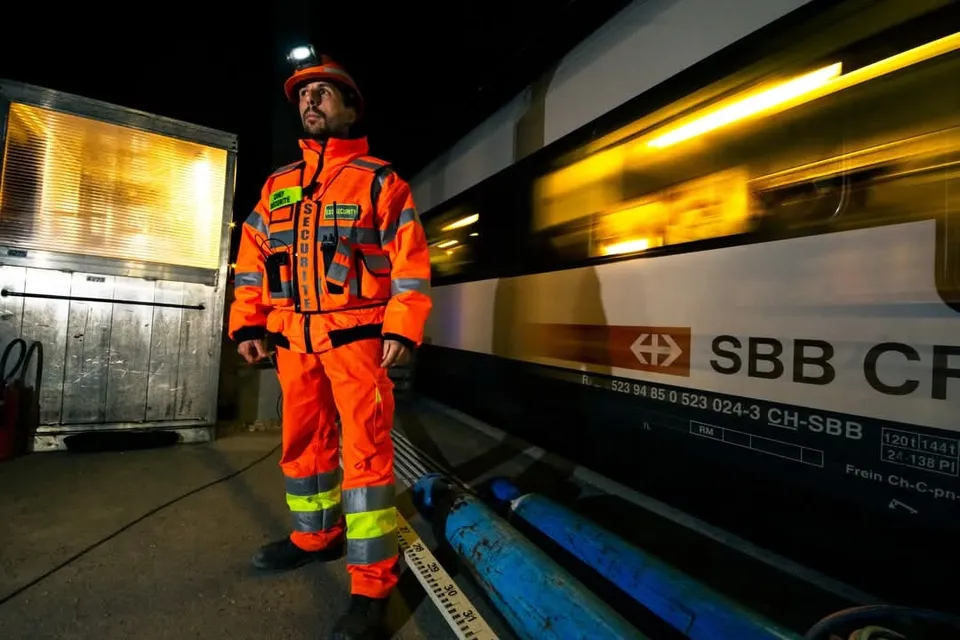 Chef sécurité en tenue orange sur un quai, de nuit, à côté d’un train CFF en passage